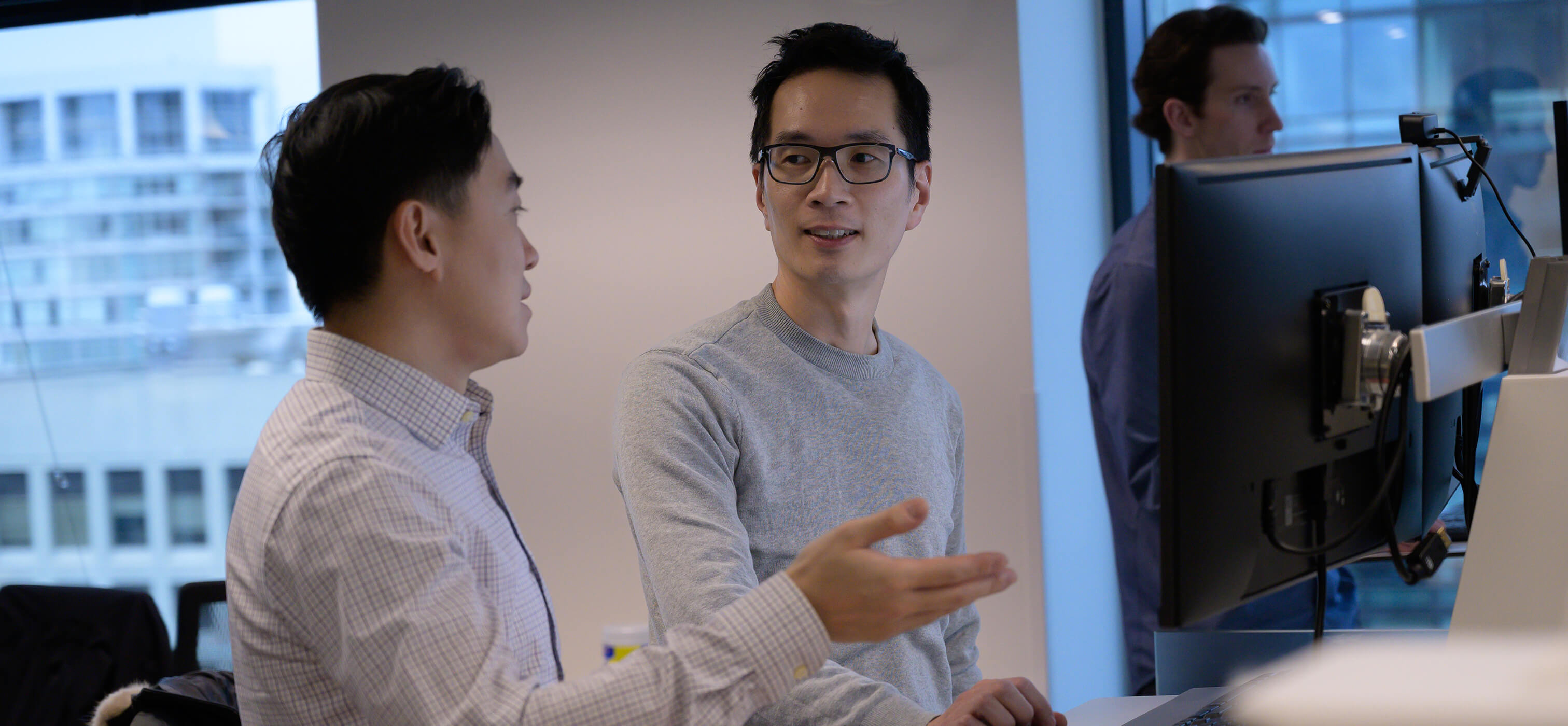 Two members of the Q Team standinga their workstations and speaking to each other.